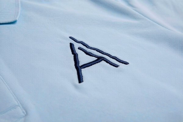 Fine embroidered corporate logo patch on a light blue company polo shirt chest, crisp stitching detail